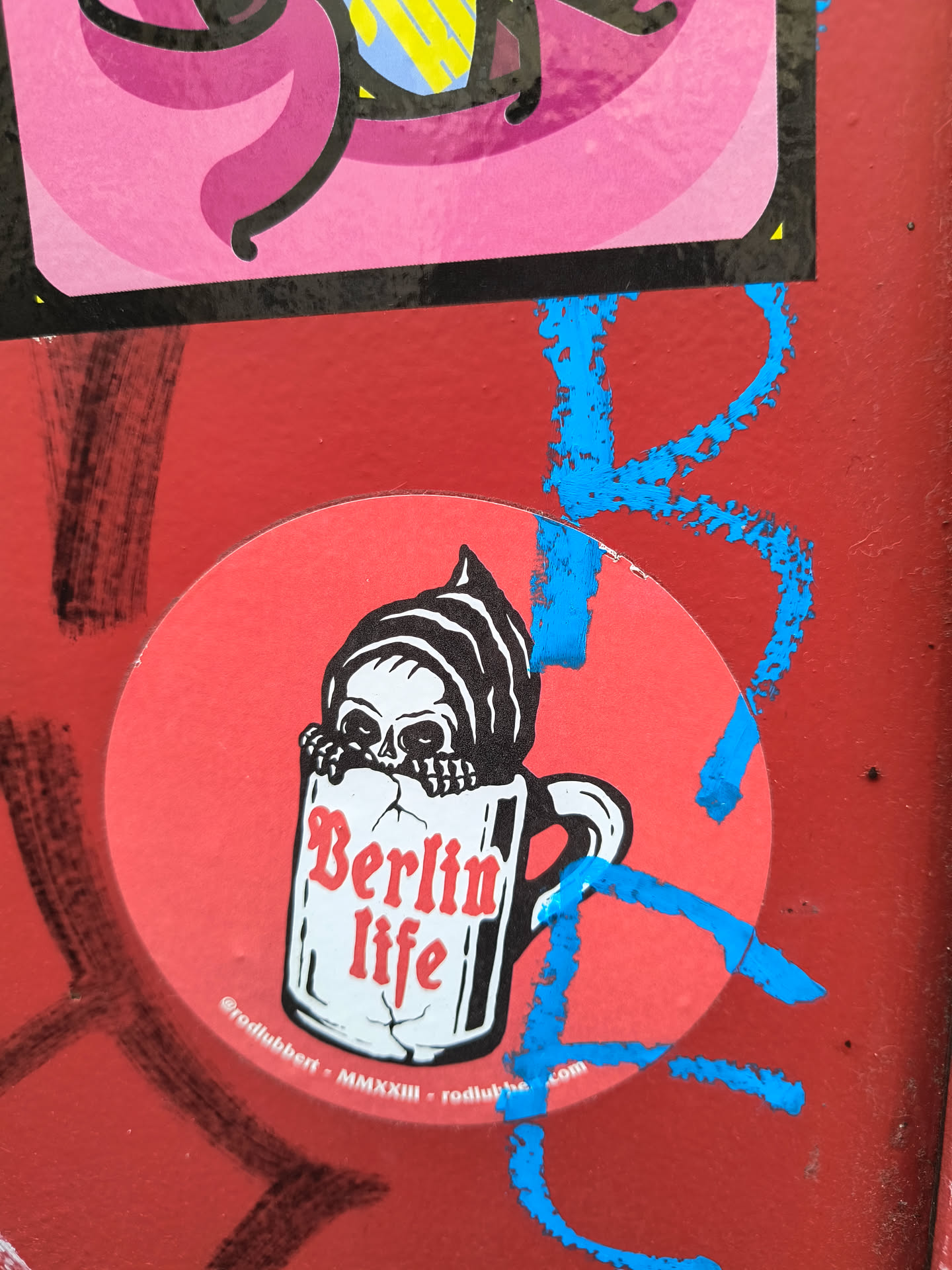 A round sticker with a red background features a Grim Reaper peering out from a mug that reads "Berlin Life." The sticker also displays social media handles.