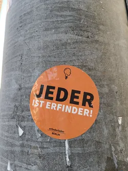 An orange round sticker with the text "Jeder ist Erfinder!" in black bold letters. At the bottom, it says "erfinderladen BERLIN" in a smaller font.  A small logo depicting a light bulb with a head is present at the top. The sticker is affixed to a grey concrete surface.