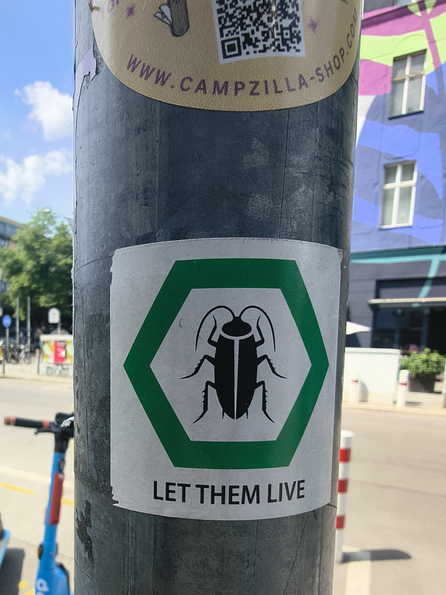 Square white sticker depicting a black cockroach silhouette inside a green hexagon. Below, the text 'LET THEM LIVE' is written in black capital letters.