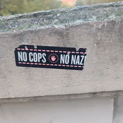 Street sticker A black and pink sticker with the text "NO COPS NO NAZIS" and a pink hand symbol in the center. The sticker is attached to a light grey wall.