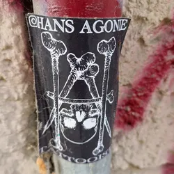 Street sticker A black and white sticker with an illustration of a skull bound by bones. The text "Hans Agone" is visible at the top. It appears to be a tattoo artist's sticker.