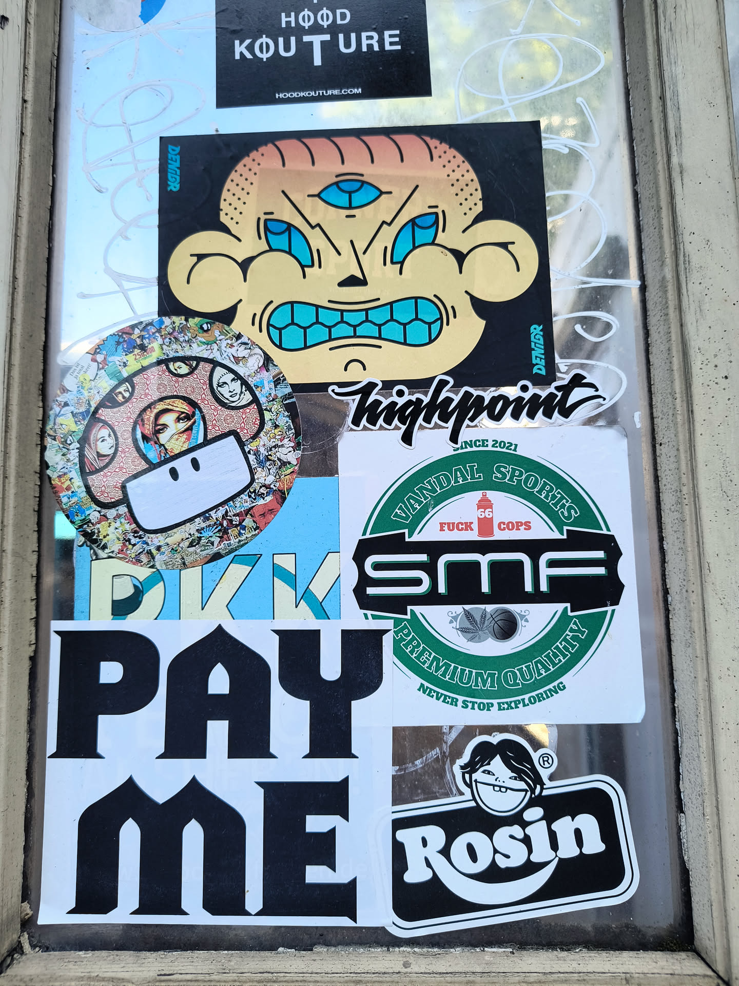 The image showcases an assortment of stickers adhered to a window. Notable designs include a three-eyed character by 'Denigr', the 'Hood Kouture' brand, and a sticker from 'Vandal Sports' with 'FUCK COPS' and 'SMF' prominently displayed. Other stickers include text 'PAY ME', a mushroom with faces, Highpoint script and Rosin character