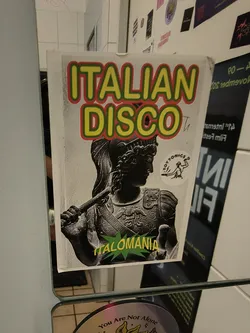 A rectangular sticker featuring a vibrant, retro-inspired design. The top half displays the words 'ITALIAN DISCO' in large, bold yellow letters with a red and green outline. Below this, a grayscale image of a classical Roman-style statue or bust, possibly a soldier or emperor, is depicted in profile, facing right and holding a staff or weapon. In the bottom left corner, a green starburst shape contains the yellow text 'ITALOMANIA'. To the right of the statue's head, a small circular white sticker with a black line drawing of a figure throwing an object and the text 'TOYTONICS' is visible. The overall background of the main sticker is a light, slightly textured white.