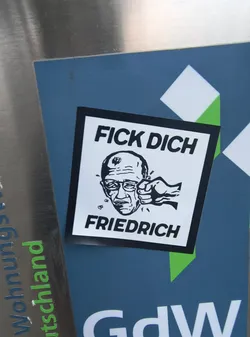 Street sticker The sticker is square with a black border. In the center, it features a black and white illustration of a man's head with glasses, being punched in the face by a fist. Above the illustration, the text 'FICK DICH' is written in bold black capital letters. Below the illustration, the name 'FRIEDRICH' is written in similar bold black capital letters.