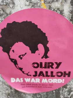 Street sticker Pink round sticker with a black silhouette of a person's head and shoulders.  Text on the sticker reads "OURY JALLOH" in large letters and "DAS WAR MORD!" in smaller letters. Below, smaller text states that Oury Jalloh was burned to death by police in January 2005. The design appears to be a memorial or protest graphic. 