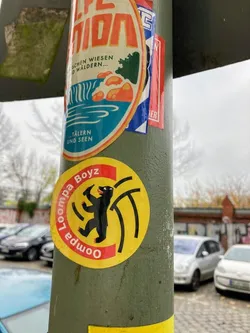 Street sticker The sticker is circular with a bright yellow background. Its main element is a stylized black bear figure, depicted in a dynamic, seemingly dancing or striding pose, with its tongue slightly extended. Behind the bear, several thick, abstract black lines radiate outwards, suggesting motion or energy. Encircling the bear is a red band where the text 'Loompa Boyz' is written in black, with 'Loompa' appearing upside down on the left curve and 'Boyz' right-side up on the right curve.