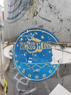 A round blue sticker with the text Zirkus Mond, a yellow moon and stars. It's partially damaged and peeling off a grey pole.