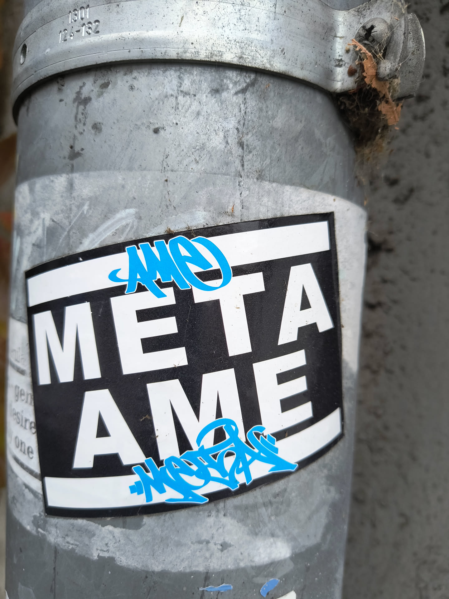 The sticker features a black rectangular background with a white border. Prominently displayed in large, white, sans-serif capital letters are the words 'META' on the top line and 'AME' on the bottom line. Above and below the main text, there are stylized blue graffiti tags. The top tag is partially obscured by the sticker's edge, while the bottom tag is more fully visible. The sticker is applied to a weathered, metallic pole.