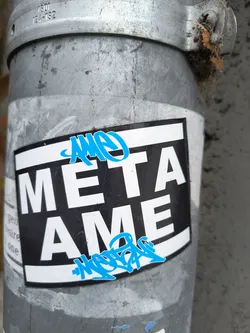 Street sticker The sticker features a black rectangular background with a white border. Prominently displayed in large, white, sans-serif capital letters are the words 'META' on the top line and 'AME' on the bottom line. Above and below the main text, there are stylized blue graffiti tags. The top tag is partially obscured by the sticker's edge, while the bottom tag is more fully visible. The sticker is applied to a weathered, metallic pole.