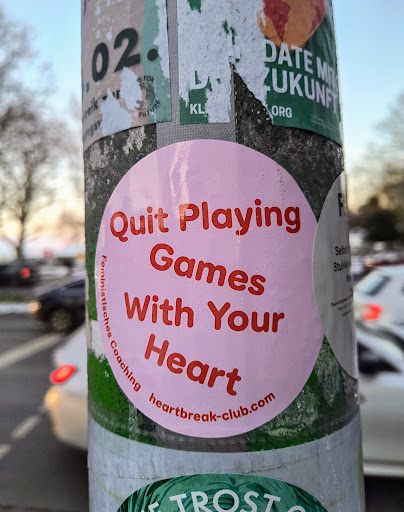 Street sticker A round, pink sticker with red text is adhered to a pole. The text reads "Quit Playing Games With Your Heart" and "Feministisches Coaching heartbreak-club.com".