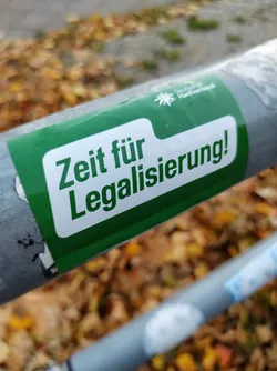 A green sticker with white text on a metal pole. The text says "Zeit für Legalisierung!" which translates to "Time for legalization!" in German.  The sticker also features a small logo. The background is blurred, showing fallen autumn leaves.