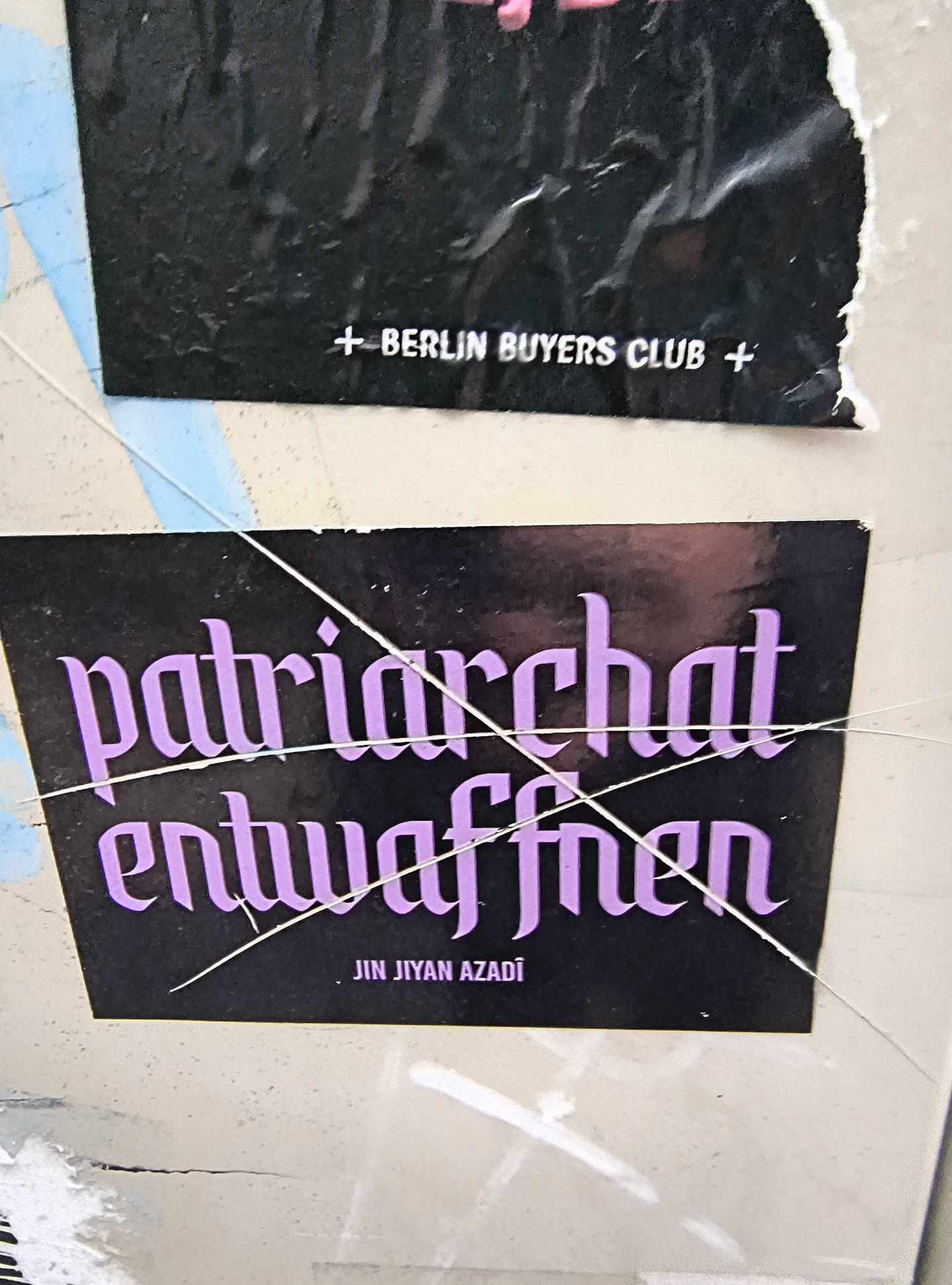 The sticker is rectangular with a black background. Prominently displayed in the center are two lines of text in a stylized, gothic-like font, colored in a vibrant lavender. The top line reads 'patriarchat' and the bottom line reads 'entwaffnen'. Below these main lines, in smaller, white, sans-serif font, is the text 'JIN JÎYAN AZADÎ'. The sticker appears to be slightly worn and has a few thin, white lines crossing over it, possibly from tape or cracks in the surface it's adhered to.
