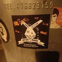 Street sticker A black sticker with a white rabbit illustration in the center. The text on the sticker says "Der Weiße Hase" which translates to "The White Rabbit" in German. It also includes the address "Revaler Str 99 | 10245 Berlin" and a Facebook link. The style is reminiscent of a techno club flyer.