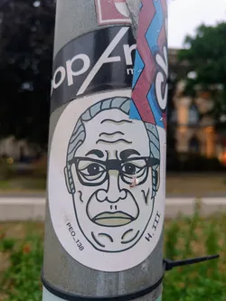 Round sticker featuring a stylized illustration of a man wearing glasses. The style is minimalistic with black outlines and muted colors.  The sticker has a small Instagram handle and number visible at the bottom. 