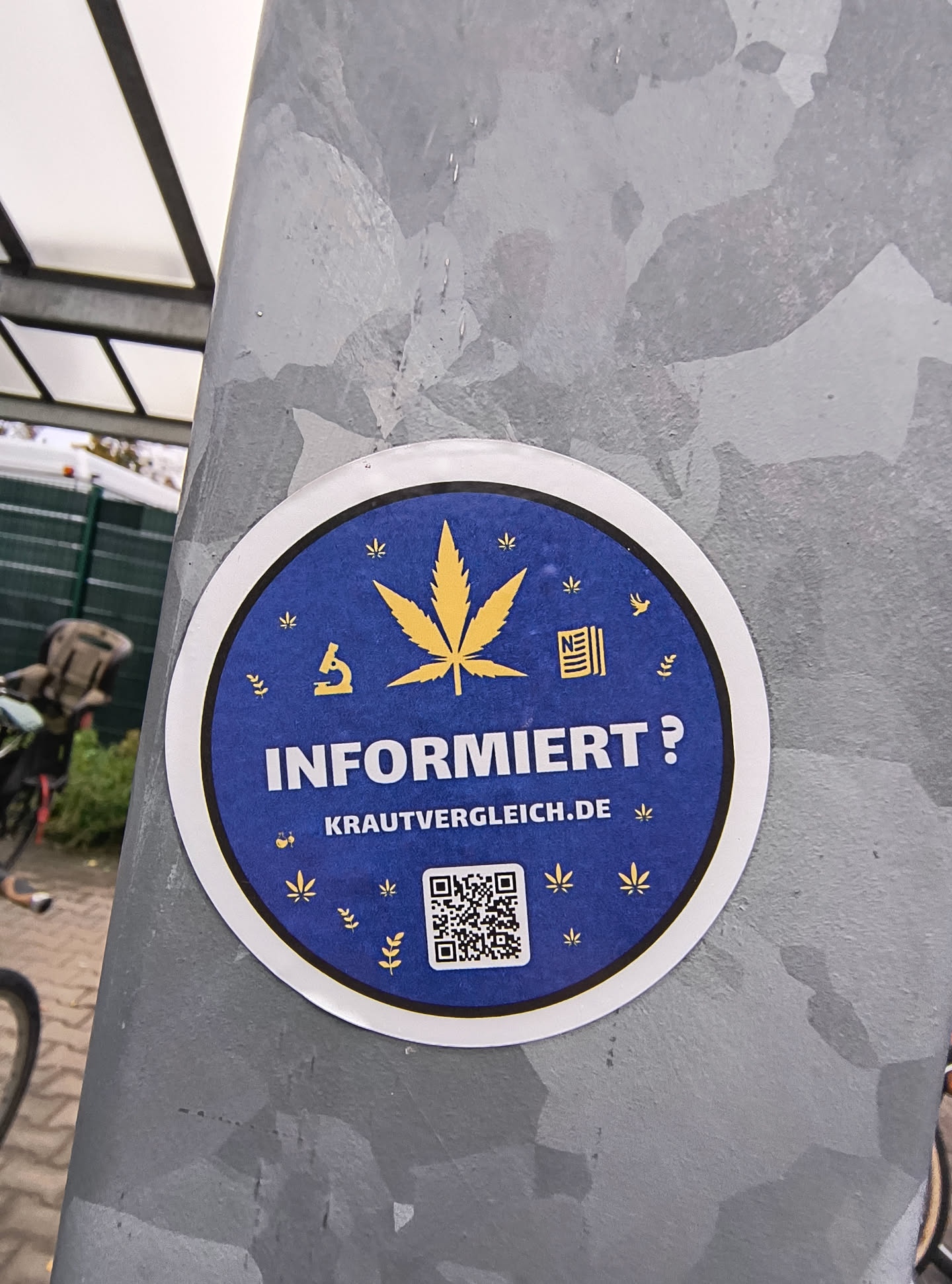 A circular sticker with a white border on a dark blue background. The center features a prominent yellow cannabis leaf. To the left of the leaf is a yellow microscope icon. To the right of the leaf is a yellow icon resembling stacked papers or a book. Scattered around the central elements are smaller yellow cannabis leaf outlines and sprigs. The text 'INFORMIERT?' is displayed in large white capital letters below the cannabis leaf, followed by 'KRAUTVERGLEICH.DE' in smaller white letters. At the bottom center is a QR code.