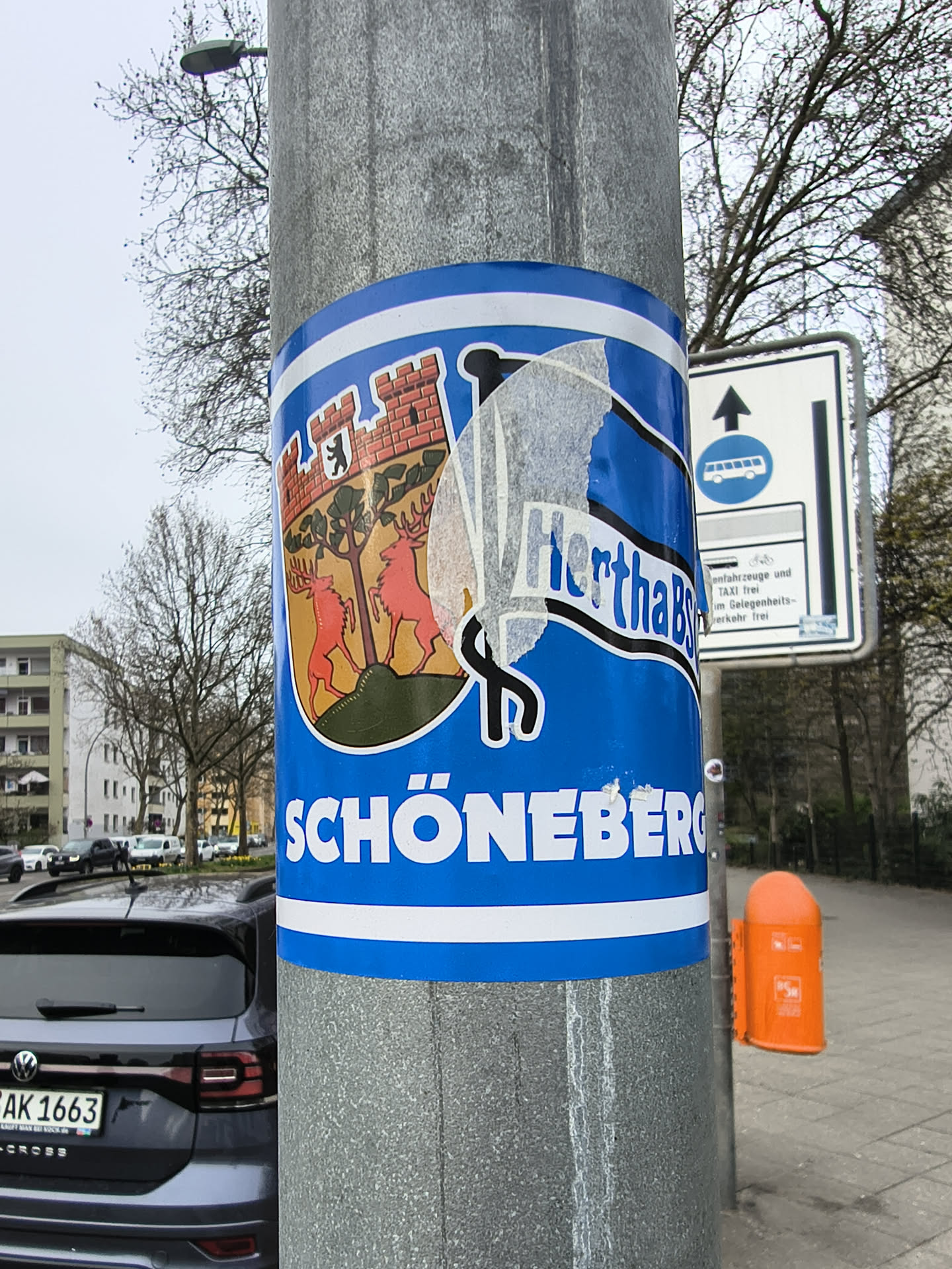 A rectangular sticker with rounded corners, predominantly blue with white horizontal stripes at the top and bottom. The left side features the coat of arms of Schöneberg, depicting a red brick castle with a black bear on its wall, and two red deer standing on a green hill against a golden-yellow background. The right side contains partially peeled white text with a black outline, which appears to read 'HerthaBSC'. Below this, in large, bold white capital letters, is the word 'SCHÖNEBERG'. The sticker shows signs of wear and peeling, particularly over the 'HerthaBSC' section.