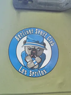 Street sticker A circular sticker featuring a cartoonish bear character as its central motif. The bear has grey fur, a dark snout, and piercing blue eyes, wearing a light blue fedora hat with a white band and a matching light blue shirt. Its arms are crossed, and it holds two small, stylized bottles (possibly vapes or miniature drinks) in its paws. The bear has a determined or slightly grumpy expression. The central image is set against a white background, encircled by a thick blue band. Along the top curve of this blue band, the words "Berliner Sport Club" are written in white, stylized letters, and along the bottom curve, "Los Spritos" is written in similar white lettering.