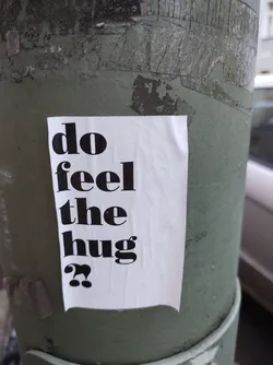 Street sticker A white sticker with black text on a green pole. The text says: "do feel the hug??"
