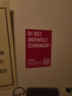 Street sticker A rectangular sticker with a bright red background and white text. The text at the top asks "DU BIST UNGEWOLLT SCHWANGER?" (Are you unintentionally pregnant?). Below this, in smaller white text, is a warning: "VORSICHT! Das passiert, wenn die AfD das Sagen hat →" (Caution! This happens when the AfD is in charge →). To the right of this text is a QR code. Below the QR code and warning, in very small print, is a website address: "www.aufstehen-gegen-rassismus.de/faktencheck". The sticker appears to be adhered to a wall.