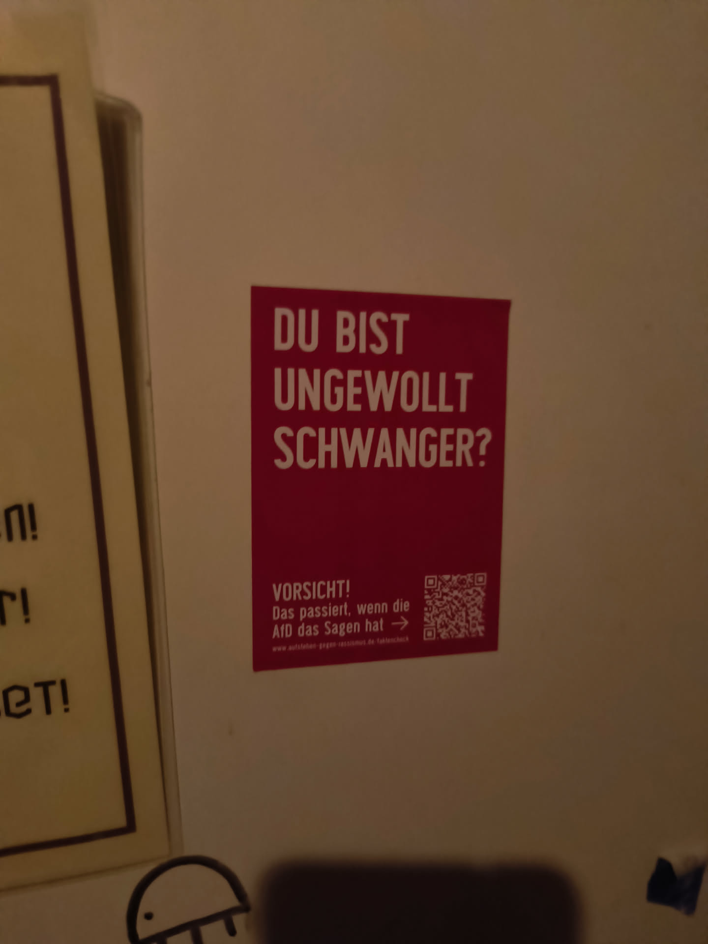 A rectangular sticker with a bright red background and white text. The text at the top asks "DU BIST UNGEWOLLT SCHWANGER?" (Are you unintentionally pregnant?). Below this, in smaller white text, is a warning: "VORSICHT! Das passiert, wenn die AfD das Sagen hat →" (Caution! This happens when the AfD is in charge →). To the right of this text is a QR code. Below the QR code and warning, in very small print, is a website address: "www.aufstehen-gegen-rassismus.de/faktencheck". The sticker appears to be adhered to a wall.