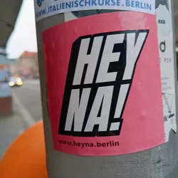 Pink and black sticker with the text 'Hey Na!' and the website www.heyNa.berlin. The sticker is placed on a grey pole.