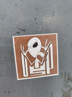 Brown square sticker with a cartoon alien sticking its tongue out. The alien is winking and making a funny face. There are some lines behind the alien that look like a maze or a code. The sticker is on a grey surface.