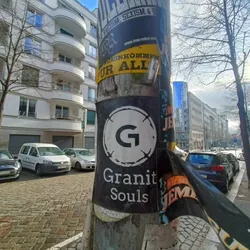 Black and white sticker with a circle and the letter G in the center. Below it, the text Granit Souls is written. The sticker is attached to a pole with other posters and stickers.