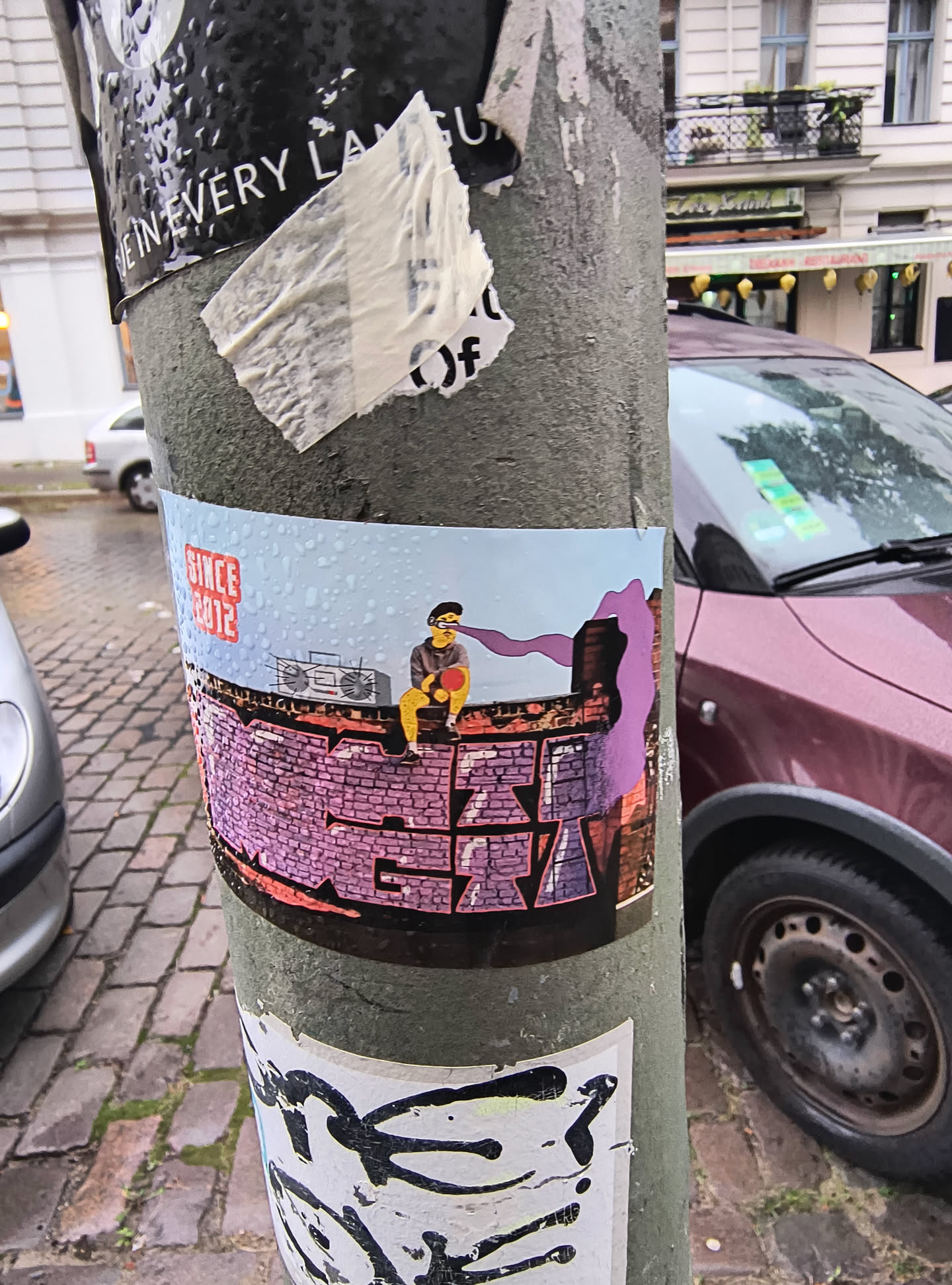 The sticker features a light blue background with water droplets, suggesting a rainy or wet environment. In the center, a cartoon character wearing a yellow hoodie and black beanie sits on a brick wall. The character is holding a red object, possibly a ball or a record, and has purple smoke or energy emanating from their eyes. Below the character, the brick wall is depicted with graffiti that spells out a word in purple and orange. To the left of the character, a vintage boombox is illustrated. The text 'SINCE 2012' is prominently displayed in a red, blocky font in the upper left corner.