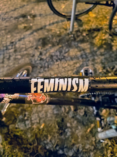 Close-up of a black bicycle frame with a 'Feminism' sticker on it. The sticker is white with black lettering and a black border. The photo is taken outdoors, and the background is blurry, with some ground and other parts of the bike visible.