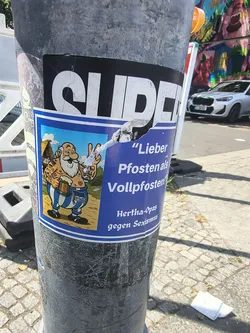 Blue sticker featuring a cartoon of a tattooed, beer-holding elderly man in blue-and-white striped pants. German text translates to 'Better a post than an idiot!' and 'Hertha-Grandpas against Sexism'.