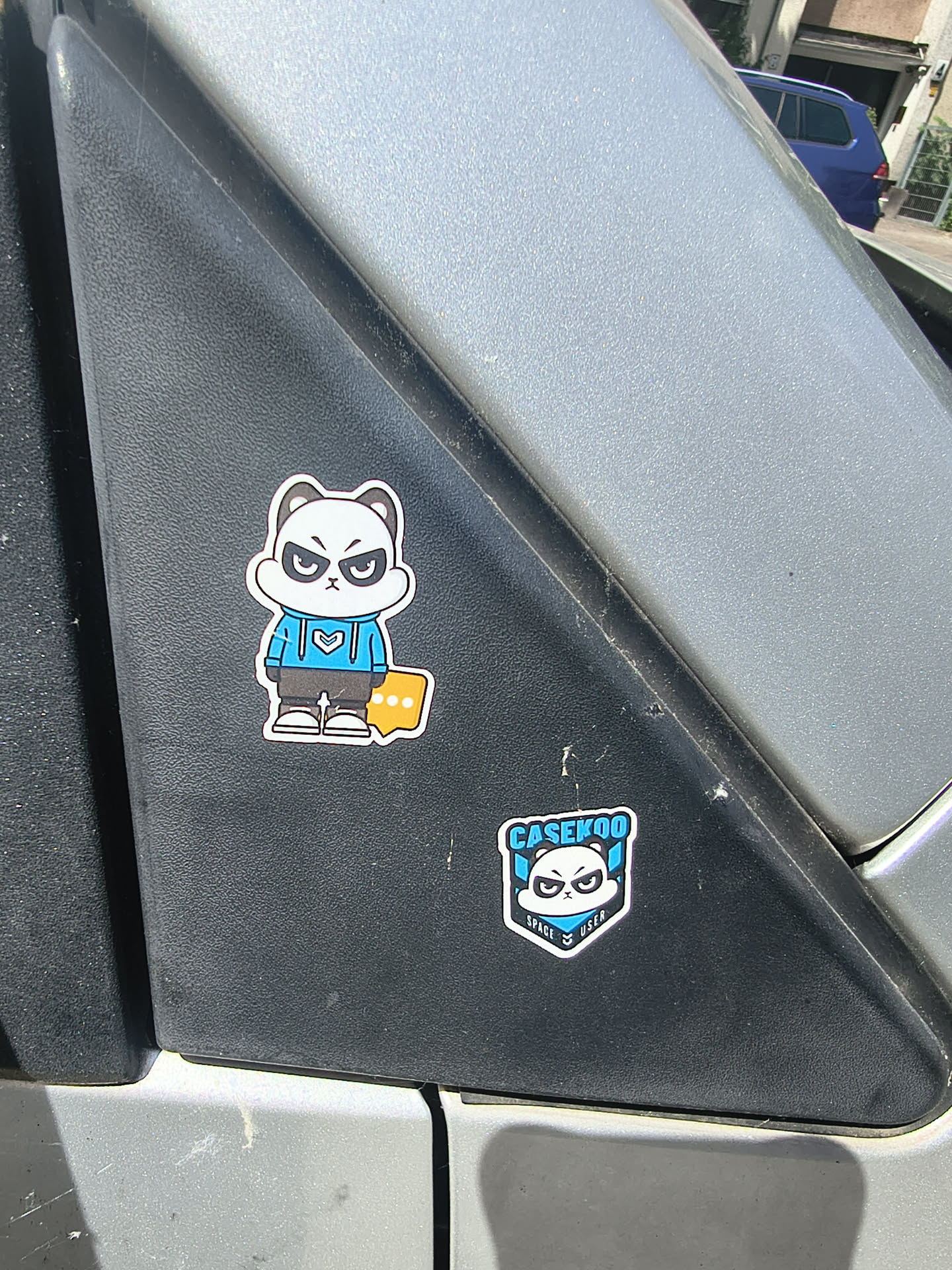 Two die-cut stickers on black plastic. The upper sticker depicts a full-body cartoon panda with an angry expression, wearing a blue hoodie and grey pants. The lower sticker is a shield-shaped logo featuring the panda's face, branded with 'CASEKOO' and 'SPACE USER'.