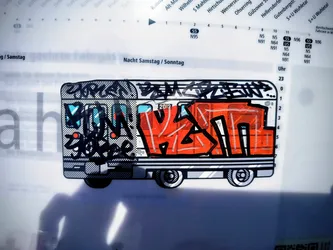 Street sticker The sticker features a stylized depiction of a train car, rendered in a graffiti art style. The main body of the train is predominantly bright orange with thick black outlines and white highlights. The front of the train has a black and white patterned section with blue accents and some red circular elements. Above the orange section, there is black graffiti lettering with a dotted texture. The wheels are black and white, with a circular, gear-like design. The overall aesthetic is bold, colorful, and reminiscent of street art.