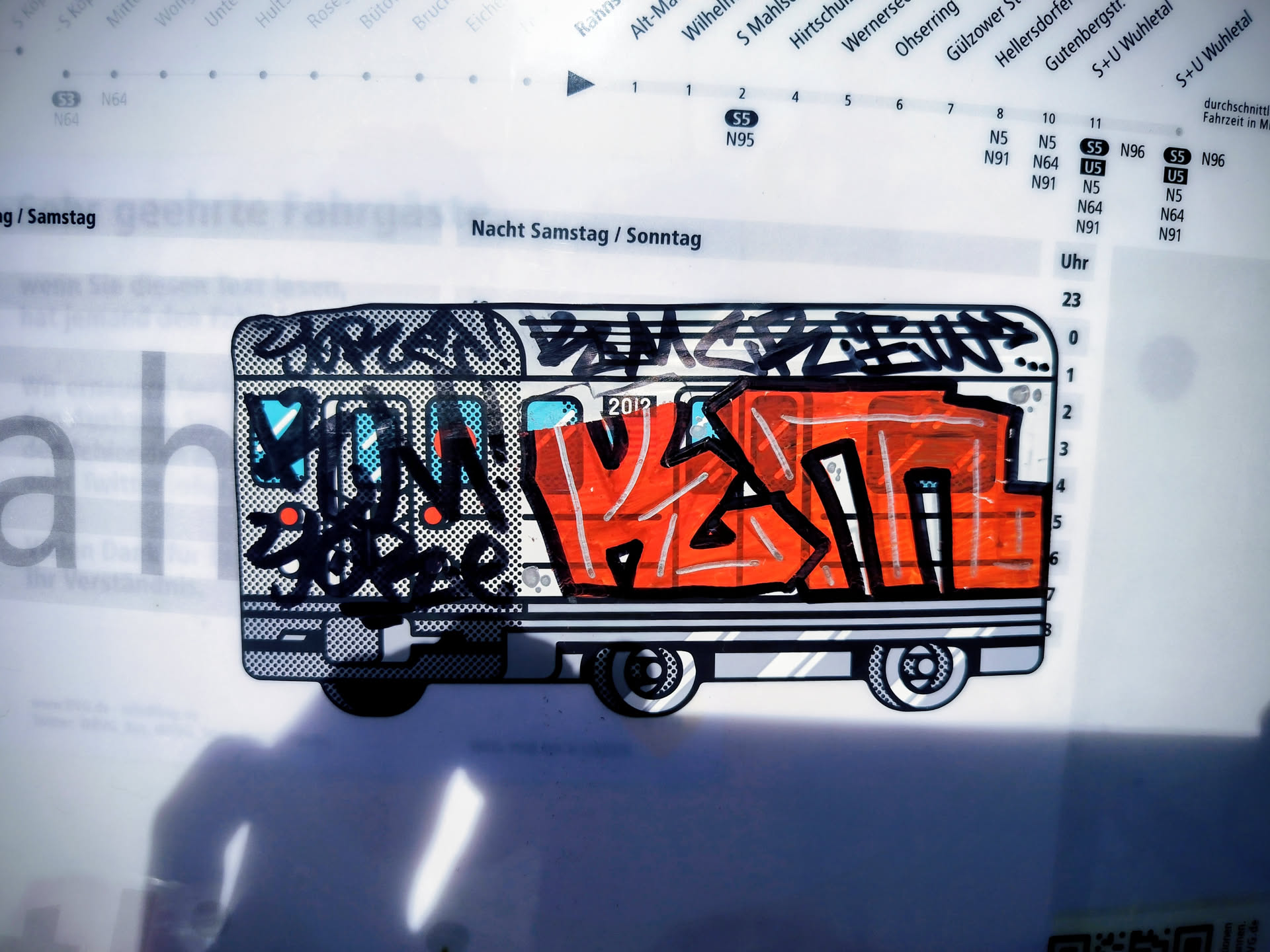 The sticker features a stylized depiction of a train car, rendered in a graffiti art style. The main body of the train is predominantly bright orange with thick black outlines and white highlights. The front of the train has a black and white patterned section with blue accents and some red circular elements. Above the orange section, there is black graffiti lettering with a dotted texture. The wheels are black and white, with a circular, gear-like design. The overall aesthetic is bold, colorful, and reminiscent of street art.