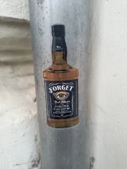 Street sticker The sticker is shaped like a classic whiskey bottle, predominantly brown for the liquid and black for the label and bottle cap. The label features the word 'FORGET' in a large, stylized white font at the top. Below it, a prominent eye with a blue iris and an orange/yellow glow around it is depicted. Underneath the eye, smaller white text reads 'But Never EVER FORGIVE,' followed by '3 Litre 100% Vol.' and 'RAGE, REVENGE & SELF-DESTRUCTION.' The bottle cap area has 'No. REGRETS.' in white text. Thin white text lines run vertically along the sides of the main label, reading 'LET IT GO' on the left and 'LET THE PAST BE THE PAST' on the right. The sticker has a clear border, suggesting it's cut out precisely to the bottle's shape and adhered to a textured, grey metallic pipe.