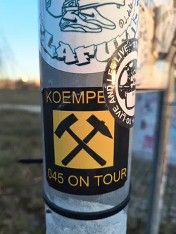 Street sticker A rectangular sticker with a black background and yellow text and graphics. At the top, the word "KOEMPE" is written in yellow sans-serif capital letters. Below this, the central graphic features two stylized black hammers crossed over a bright yellow square background. Below the yellow square, the text "045 ON TOUR" is displayed in yellow capital letters. The sticker shows minor wear, including some scratches and a slight tear at the top left corner.