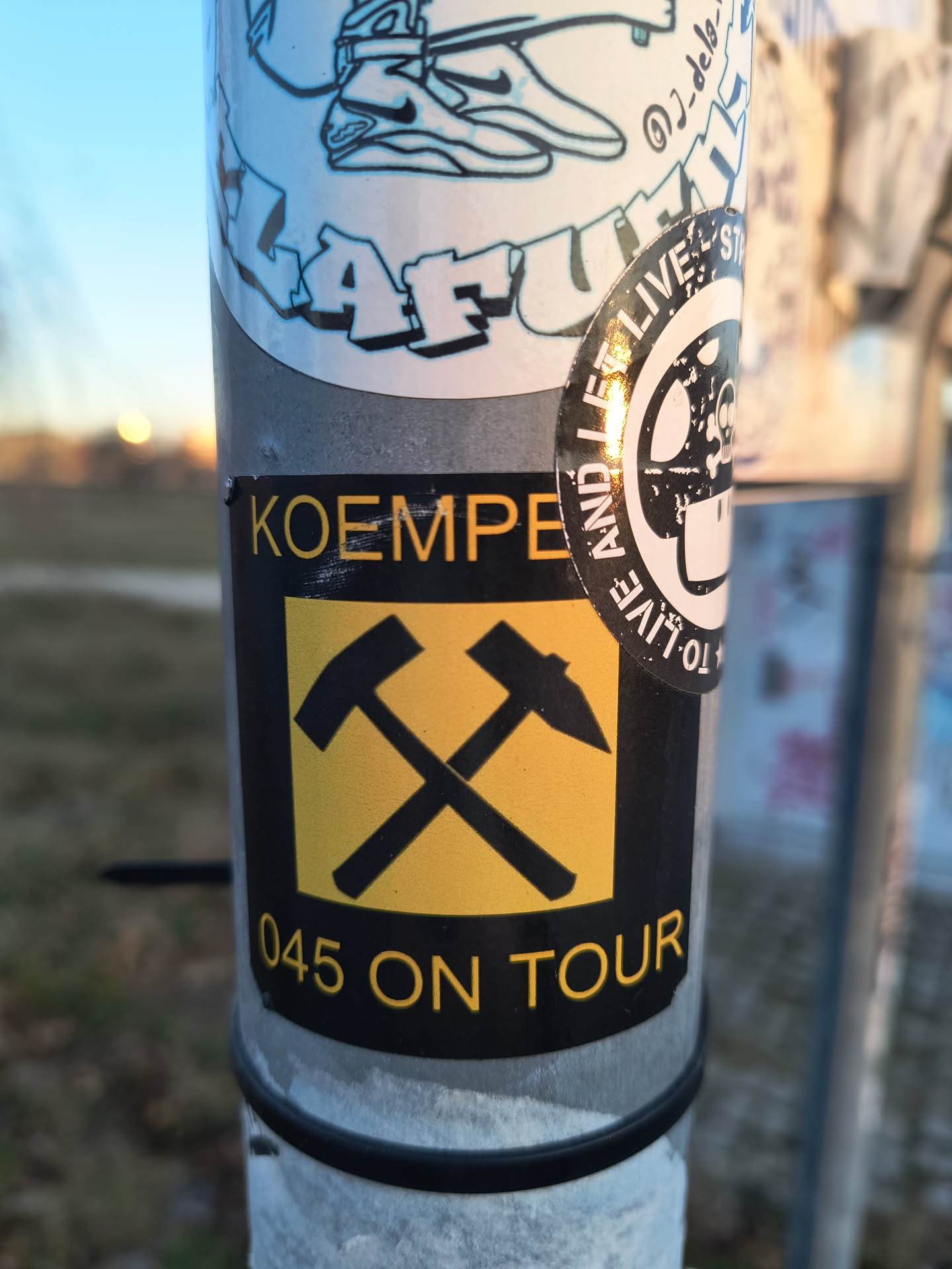Street sticker A rectangular sticker with a black background and yellow text and graphics. At the top, the word "KOEMPE" is written in yellow sans-serif capital letters. Below this, the central graphic features two stylized black hammers crossed over a bright yellow square background. Below the yellow square, the text "045 ON TOUR" is displayed in yellow capital letters. The sticker shows minor wear, including some scratches and a slight tear at the top left corner.