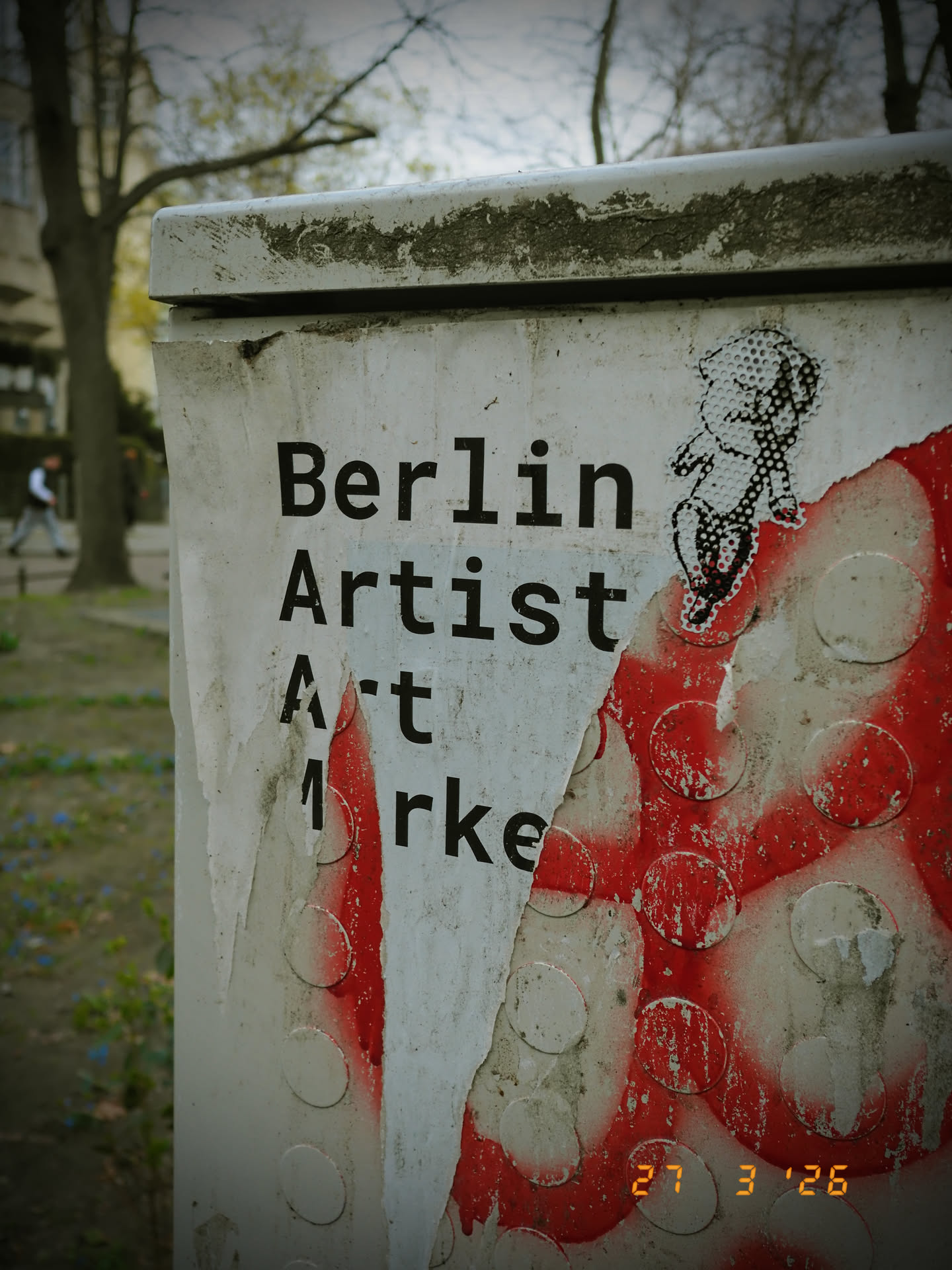 The central element is a weathered white paper sticker adhered to a dirty utility box. The sticker features bold black sans-serif text arranged vertically: 'Berlin', 'Artist', 'Art', and partially visible 'M rke' (likely 'Market'). To the upper right of the text, a small, stylized figure, possibly a child or cherub, is depicted in a black-and-white halftone or dotted pattern. This figure appears to be in motion, with its arms outstretched. A significant portion of the sticker on the lower right has peeled away, revealing a vibrant red spray-painted surface underneath, adorned with numerous lighter circular stenciled patterns. The overall appearance suggests an urban art piece that has been exposed to the elements.