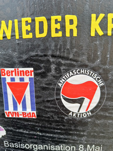 Street sticker A sticker featuring a red and black flag symbol, often associated with anti-fascist movements.  The text around the symbol says "Antifaschistische Aktion".  There is also a smaller sticker visible featuring the logo for Berliner VVN-BdA.