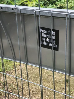Street sticker A square black sticker featuring bold white German text. It is adhered to a metallic grey sign behind a metal wire fence.