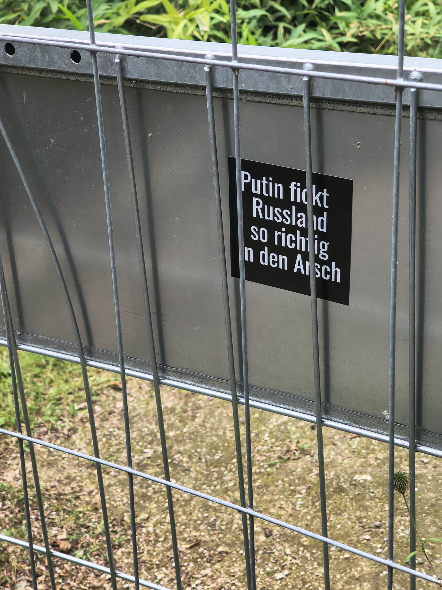 A square black sticker featuring bold white German text. It is adhered to a metallic grey sign behind a metal wire fence.
