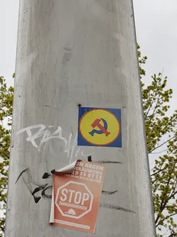 A round sticker with a yellow background and a blue border shows a hammer and sickle symbol.  It is affixed to a gray pole alongside another sticker that reads “STOP ZWANGSRAUMUNGEN” and contains phone numbers and dates. There is graffiti on the pole as well.