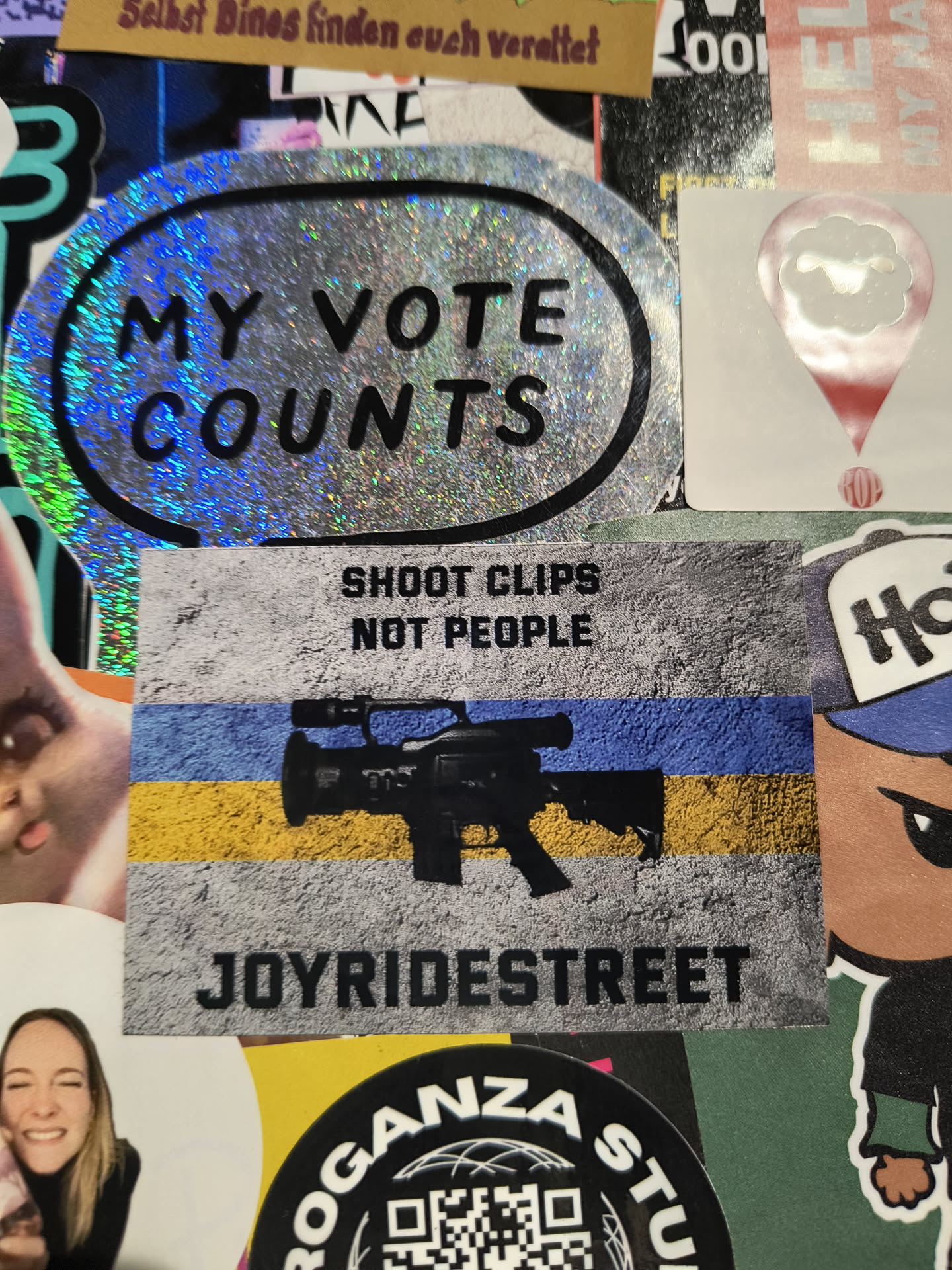 Street sticker The central sticker is a rectangular sticker with a rough, textured grey background at the top, which transitions into a flag-like pattern of blue and yellow stripes at the bottom. Superimposed on the grey area, in bold black sans-serif font, are the words 'SHOOT CLIPS NOT PEOPLE'. Below this, on the blue and yellow stripes, is an image of a professional video camera with a lens and what appears to be a gun sight mounted on top. The bottom of the sticker has the text 'JOYRIDESTREET' in bold black sans-serif font, also on a textured grey background. The sticker is surrounded by other stickers and appears to be part of a collage.