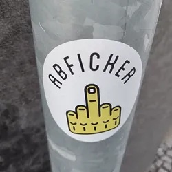 A round sticker with the word "ABFICKER" and a middle finger in yellow. The sticker is on a grey metal pole.