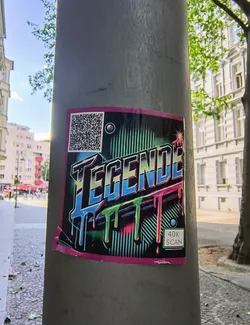 A sticker featuring the word 'TEGENDE' in a stylized, dripping font is affixed to a gray pole. The word is rendered in shades of blue and black, with the dripping effect highlighted in green and pink. The sticker has a black background with white stripes. A QR code sits in the top left corner, and the text '40K SCAN' is visible in the bottom right corner. The sticker has a pink border.
