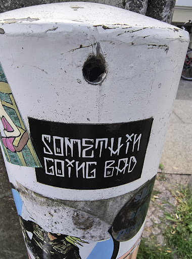 A black rectangular sticker with white text affixed to a white pillar. The text reads "SOMETHIN' GOING BAD" in a stylized, slightly distressed font. Other stickers are partially visible around it.