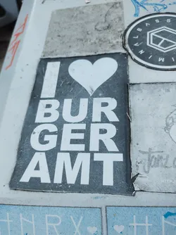Street sticker A sticker with a simple design. It says I love Burgeramt with a heart in the middle. The sticker is slightly worn and dirty, showing some signs of age and exposure to the elements. It's pasted on a wall or surface that has other stickers and graffiti around it.