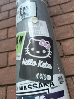 Street sticker The central sticker features a stylized black and white depiction of Hello Kitty, complete with her signature pink bow. Her eyes are depicted as spirals, and her mouth is a simple line. Below the character, the text 'Hello Keta' is written in a white, cursive font. Beneath this, smaller text reads 'auto 3INS' followed by an Instagram logo. The sticker has a dark background and appears to be applied to a metal pole, with other stickers partially visible around it.