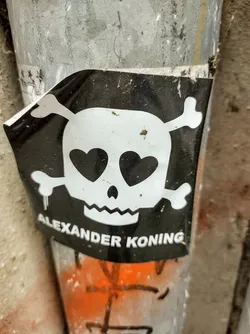 Street sticker Black and white sticker featuring a skull with hearts for eyes and crossbones. The artist's name, Alexander Koning, is printed below the skull. The sticker is affixed to a metal pole.