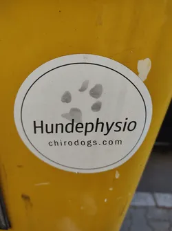 Street sticker A round white sticker on a yellow pole with a paw print and the text "Hundephysio chirodogs.com".