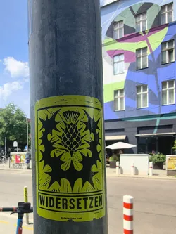 Street sticker A rectangular lime-green sticker on a pole. It displays a black, linocut-style graphic of a thistle. The bottom features the bold text 'WIDERSETZEN' and the website 'widersetzen.com' in small print.